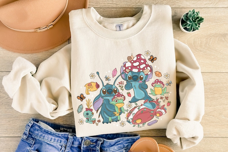 May include: A cream-colored sweatshirt with a graphic print of Stitch from Lilo & Stitch. The graphic features Stitch in various poses with mushrooms, flowers, and butterflies. The text "Stitch" is printed on the sweatshirt.