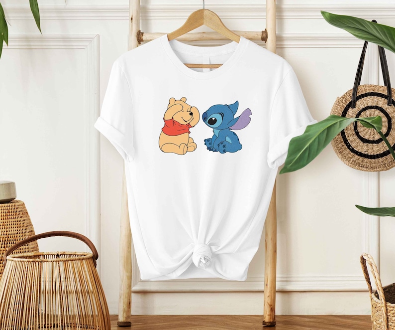 Puede incluir: Camiseta blanca con una imagen de dibujos animados de Winnie the Pooh y Stitch. Winnie the Pooh lleva una camisa roja y Stitch es azul.