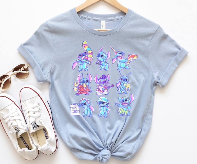 K&ouml;nnte beinhalten: Hellblaues T-Shirt mit einem farbenfrohen Aufdruck der Figur Stitch in verschiedenen Posen. Die Zeichentrickfigur wird mit verschiedenen Accessoires und Outfits dargestellt. Das T-Shirt wird auf einem Holzb&uuml;gel mit wei&szlig;en Turnschuhen und Sonnenbrillen pr&auml;sentiert.