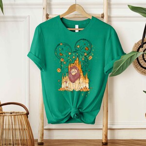May include: Green t-shirt with a graphic print of a lion in front of a castle with fireworks and a Mickey Mouse ear silhouette. The graphic is in shades of yellow, orange, and brown.