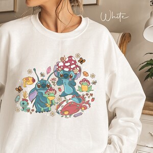 May include: White sweatshirt with a colorful graphic print of Stitch from Lilo & Stitch. The graphic features Stitch holding a frog, surrounded by mushrooms, flowers, and butterflies.