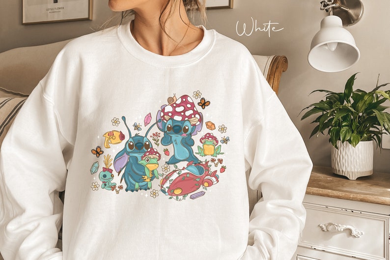 May include: White sweatshirt with a colorful graphic print of Stitch from Lilo & Stitch. The graphic features Stitch holding a frog, surrounded by mushrooms, flowers, and butterflies.