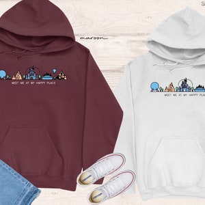 Op de afbeelding: Twee hoodies met een afbeelding van de Disney World skyline en de tekst "Meet me at my happy place". De ene hoodie is maroon en de andere is wit.
