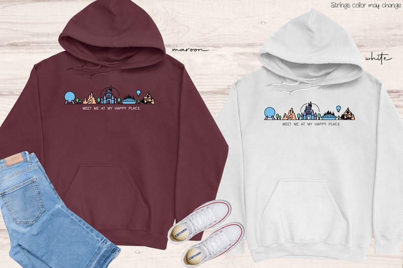 Op de afbeelding: Twee hoodies met een afbeelding van de Disney World skyline en de tekst "Meet me at my happy place". De ene hoodie is maroon en de andere is wit.