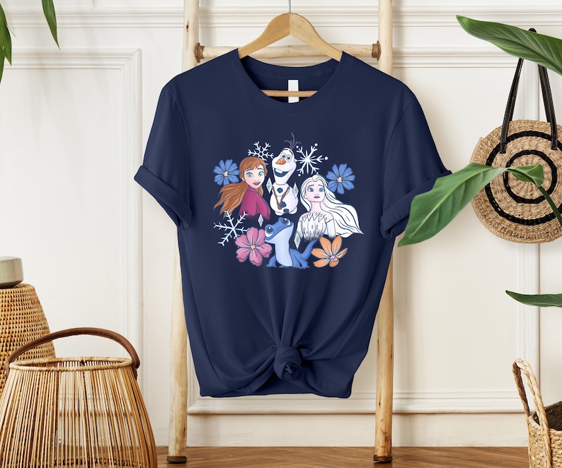 May include: A navy blue t-shirt with a graphic print of Anna, Elsa, Olaf, and Sven from the movie Frozen. The graphic is surrounded by snowflakes and flowers.