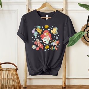 May include: A dark gray t-shirt with a colorful print of Ariel from The Little Mermaid and other characters from the movie. The print includes Ariel, Flounder, Sebastian, and a seahorse.