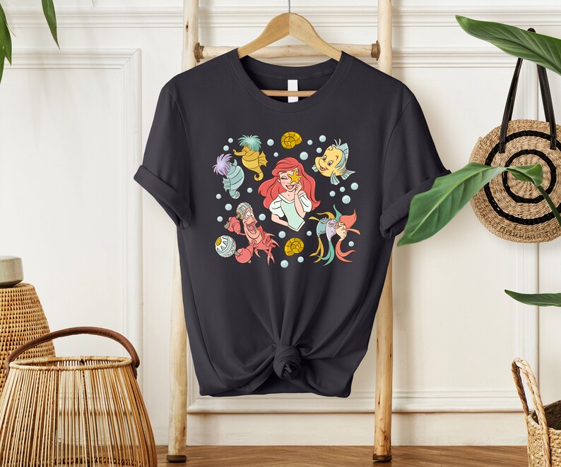 May include: A dark gray t-shirt with a colorful print of Ariel from The Little Mermaid and other characters from the movie. The print includes Ariel, Flounder, Sebastian, and a seahorse.