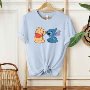 Puede incluir: Camiseta azul claro con una imagen de dibujos animados de Winnie the Pooh y Stitch. Winnie the Pooh se est&aacute; tapando los ojos con las patas y Stitch lo est&aacute; mirando con una expresi&oacute;n traviesa.