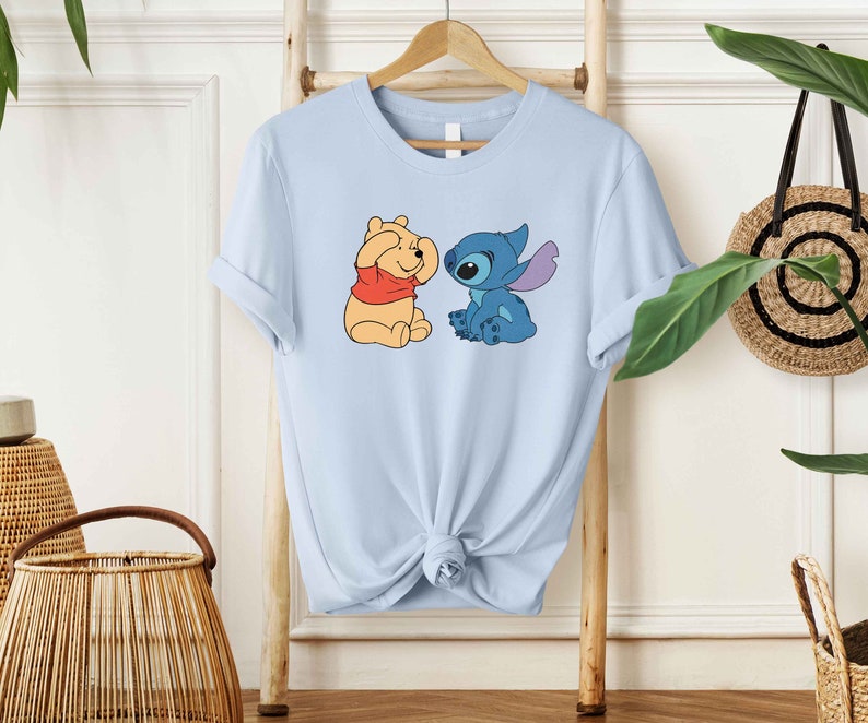 Puede incluir: Camiseta azul claro con una imagen de dibujos animados de Winnie the Pooh y Stitch. Winnie the Pooh se est&aacute; tapando los ojos con las patas y Stitch lo est&aacute; mirando con una expresi&oacute;n traviesa.