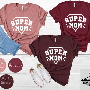 May include: Two t-shirts with the text "SUPER MOM" in a diamond shape, one in maroon and one in heather mauve. The shirts are laid out on a wooden surface with a pair of white sneakers and a pair of blue jeans.