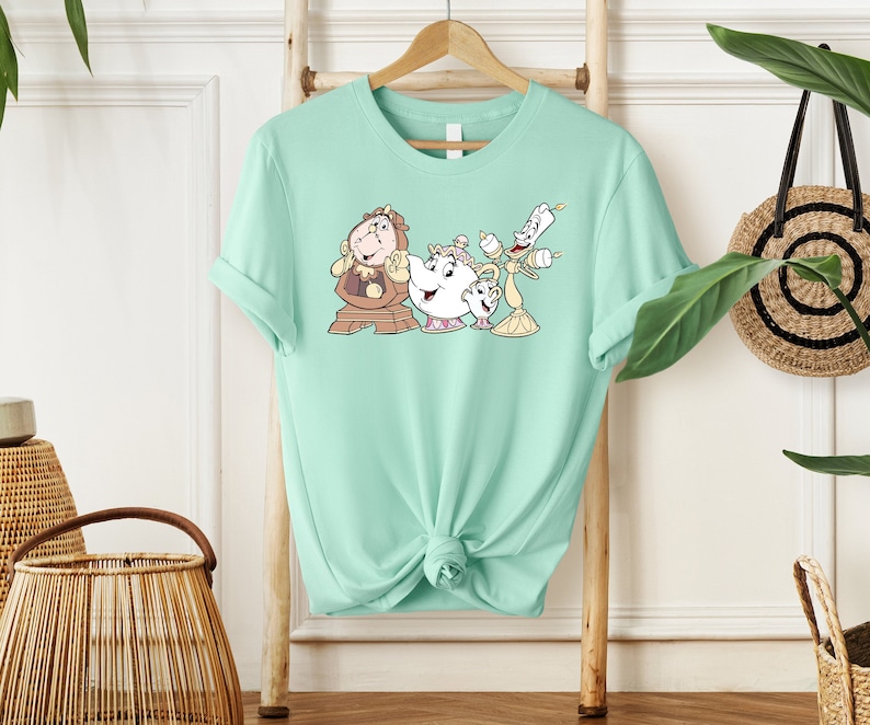 May include: A light green t-shirt with a graphic print of characters from the Disney movie Beauty and the Beast. The characters include Cogsworth, Mrs. Potts, and Lumiere.