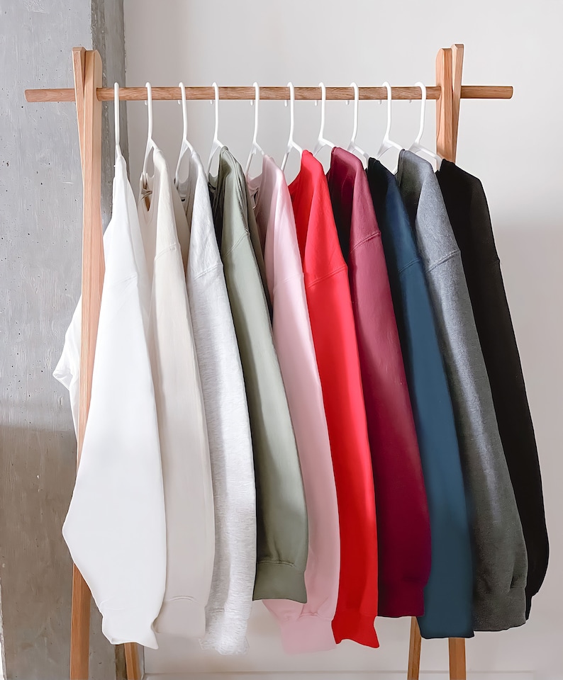 May include: A rack of 10 plain sweatshirts in various colors, including white, gray, green, pink, red, burgundy, navy, and black. The sweatshirts are hanging on white hangers.
