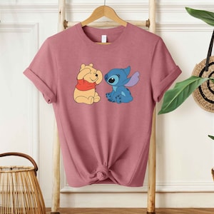 Puede incluir: Una camiseta color malva con una imagen de dibujos animados de Winnie the Pooh y Stitch. Winnie the Pooh se est&aacute; tapando los ojos con las patas y Stitch lo est&aacute; mirando con una sonrisa.