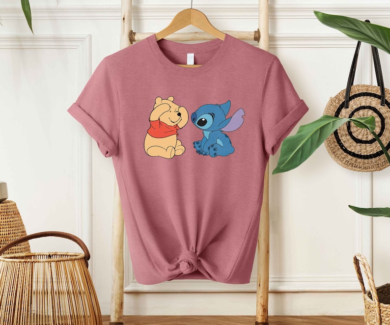 Puede incluir: Una camiseta color malva con una imagen de dibujos animados de Winnie the Pooh y Stitch. Winnie the Pooh se est&aacute; tapando los ojos con las patas y Stitch lo est&aacute; mirando con una sonrisa.