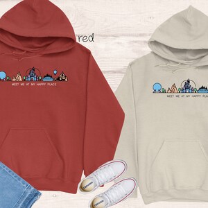 Op de afbeelding: Twee hoodies met een afbeelding van de Disney World skyline en de tekst "Meet me at my happy place". De ene hoodie is rood en de andere is zandkleurig.
