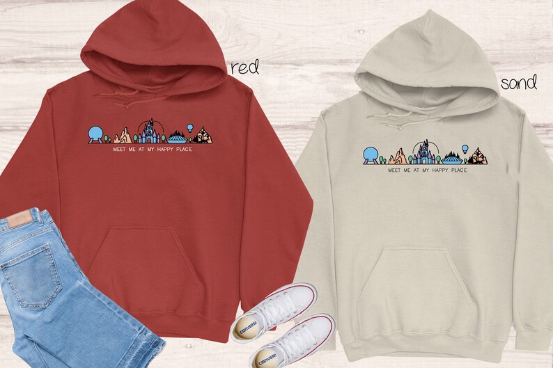 Op de afbeelding: Twee hoodies met een afbeelding van de Disney World skyline en de tekst "Meet me at my happy place". De ene hoodie is rood en de andere is zandkleurig.