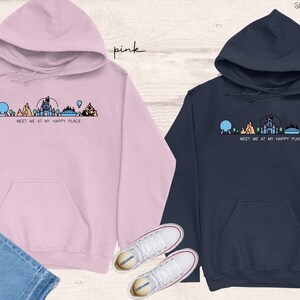 Op de afbeelding: Twee hoodies met een afbeelding van een Disney-themapark skyline en de tekst "Meet me at my happy place". Een hoodie is roze en de andere is donkerblauw.