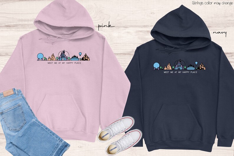 Op de afbeelding: Twee hoodies met een afbeelding van een Disney-themapark skyline en de tekst "Meet me at my happy place". Een hoodie is roze en de andere is donkerblauw.