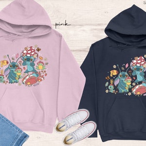 May include: Two hooded sweatshirts with a cartoon design of Stitch from Lilo & Stitch. The pink sweatshirt has a pink hood and the navy sweatshirt has a navy hood. Both sweatshirts feature a design of Stitch with mushrooms, flowers, and other nature-themed elements.
