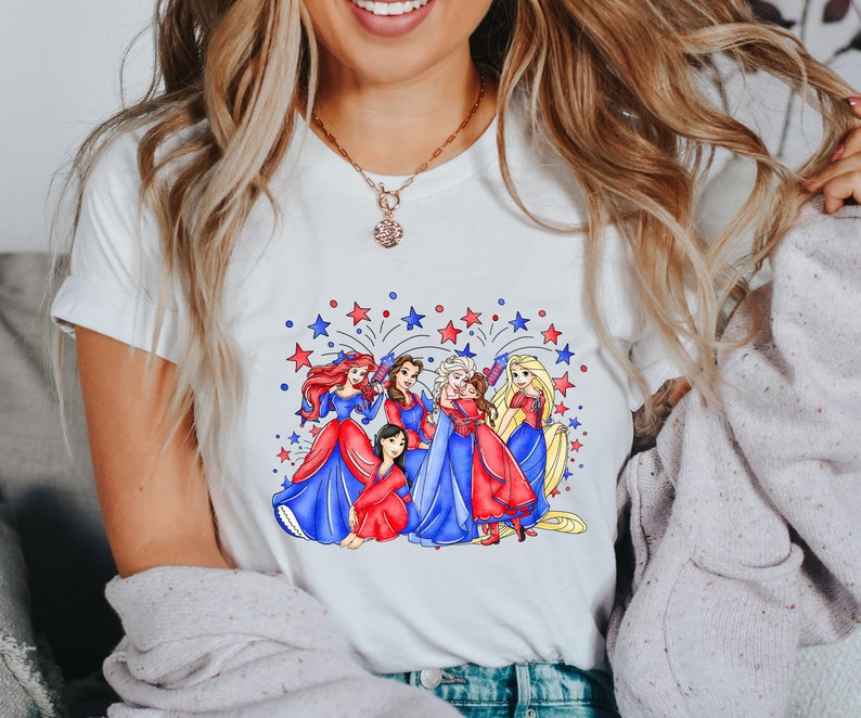 Op de afbeelding: Wit T-shirt met een afbeelding van Disney-prinsessen in rode, witte en blauwe jurken met vuurwerk op de achtergrond.