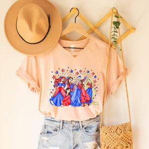 Op de afbeelding: Een perzikkleurig T-shirt met een afbeelding van Disney-prinsessen in rood, wit en blauwe jurken met vuurwerk op de achtergrond. Het shirt is gestyled met een bruine hoed, denim shorts en een geweven tas.