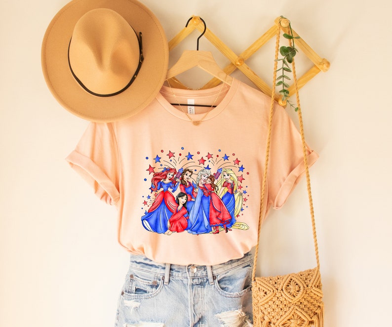 Op de afbeelding: Een perzikkleurig T-shirt met een afbeelding van Disney-prinsessen in rood, wit en blauwe jurken met vuurwerk op de achtergrond. Het shirt is gestyled met een bruine hoed, denim shorts en een geweven tas.