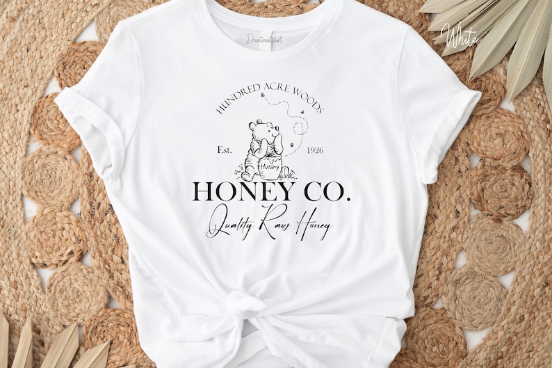 May include: White t-shirt with a black graphic design featuring Winnie the Pooh holding a jar of honey. The graphic includes the text "Hundred Acre Woods", "Est. 1926", "Honey Co.", and "Quality Raw Honey".
