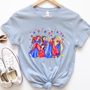 Op de afbeelding: Lichtblauw T-shirt met een afbeelding van Disney-prinsessen in rode, witte en blauwe jurken met vuurwerk op de achtergrond.