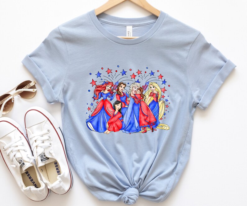 Op de afbeelding: Lichtblauw T-shirt met een afbeelding van Disney-prinsessen in rode, witte en blauwe jurken met vuurwerk op de achtergrond.