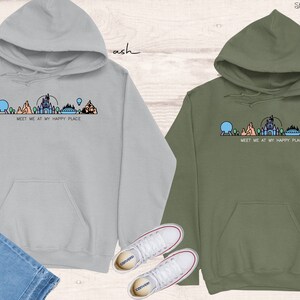 Op de afbeelding: Twee hoodies met een afbeelding van een Disney-themapark skyline en de tekst "Meet me at my happy place". De hoodie aan de linkerkant is lichtgrijs en de hoodie aan de rechterkant is legergroen.