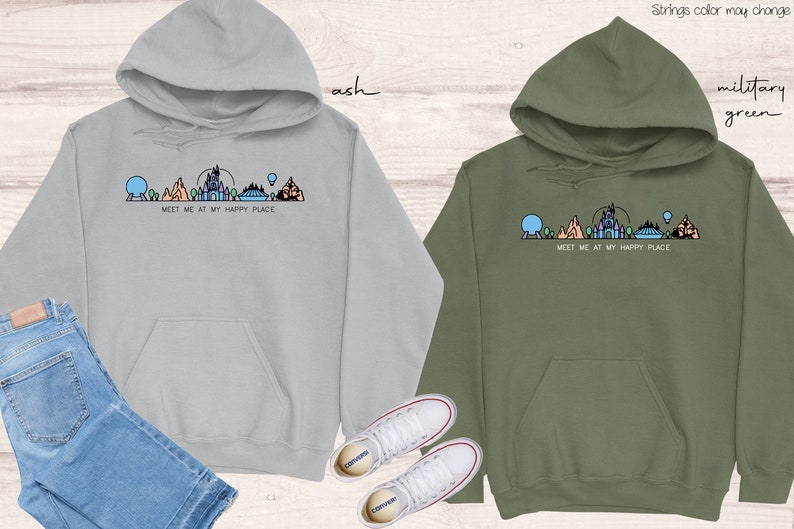 Op de afbeelding: Twee hoodies met een afbeelding van een Disney-themapark skyline en de tekst "Meet me at my happy place". De hoodie aan de linkerkant is lichtgrijs en de hoodie aan de rechterkant is legergroen.