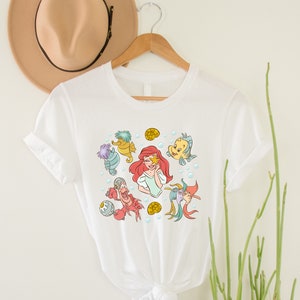 May include: White t-shirt with a colorful graphic print of Ariel from The Little Mermaid surrounded by sea creatures, including a seahorse, a crab, and a fish.