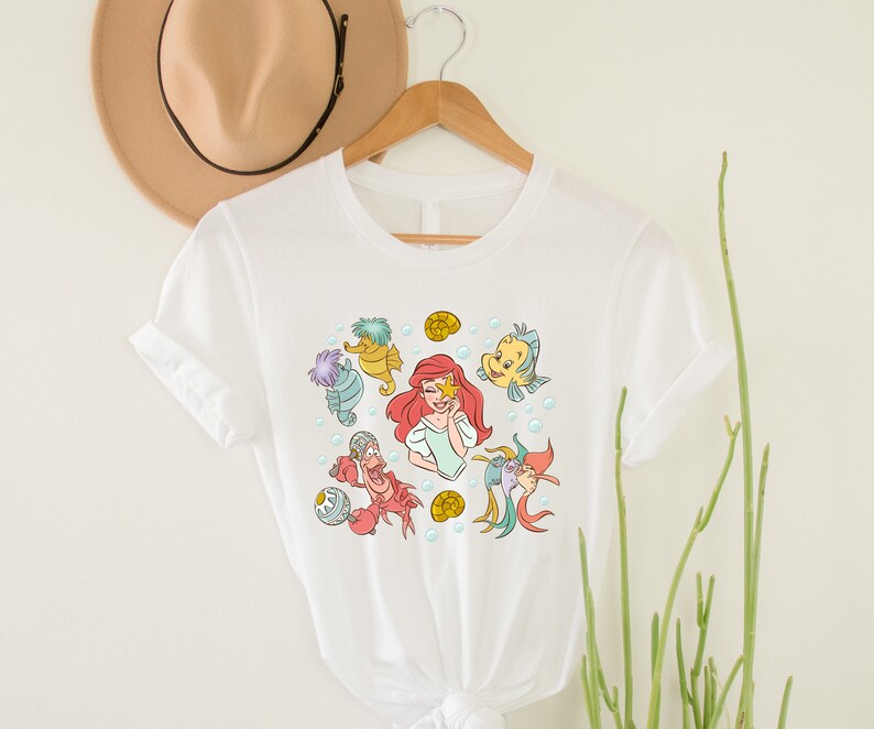 May include: White t-shirt with a colorful graphic print of Ariel from The Little Mermaid surrounded by sea creatures, including a seahorse, a crab, and a fish.