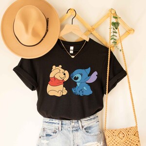 Puede incluir: Camiseta negra con un gr&aacute;fico de Winnie the Pooh y Stitch. Winnie the Pooh se est&aacute; tapando los ojos con las patas y Stitch lo est&aacute; mirando. La camiseta est&aacute; en una percha con un sombrero de paja y un bolso de paja.