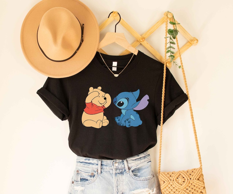 Puede incluir: Camiseta negra con un gr&aacute;fico de Winnie the Pooh y Stitch. Winnie the Pooh se est&aacute; tapando los ojos con las patas y Stitch lo est&aacute; mirando. La camiseta est&aacute; en una percha con un sombrero de paja y un bolso de paja.