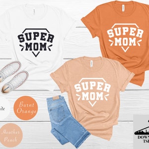 May include: Three t-shirts with the text "SUPER MOM" in a diamond shape. The shirts are white, burnt orange, and heather peach.