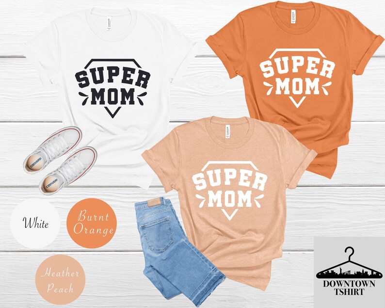May include: Three t-shirts with the text "SUPER MOM" in a diamond shape. The shirts are white, burnt orange, and heather peach.