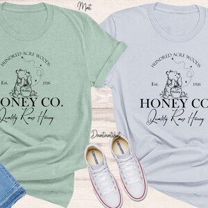 May include: Two t-shirts featuring a Winnie the Pooh inspired design. The design depicts a bear holding a jar of honey with the text "Hundred Acre Woods" and "Honey Co. Quality Raw Honey". The t-shirts are in mint and light blue.
