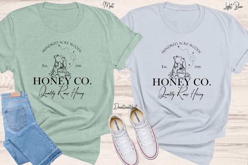 May include: Two t-shirts featuring a Winnie the Pooh inspired design. The design depicts a bear holding a jar of honey with the text "Hundred Acre Woods" and "Honey Co. Quality Raw Honey". The t-shirts are in mint and light blue.