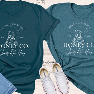 May include: Two teal blue t-shirts with a white graphic design of a bear holding a jar of honey. The graphic includes the text "Hundred Acre Woods", "Est. 1926", "Honey Co.", and "Quality Raw Honey".