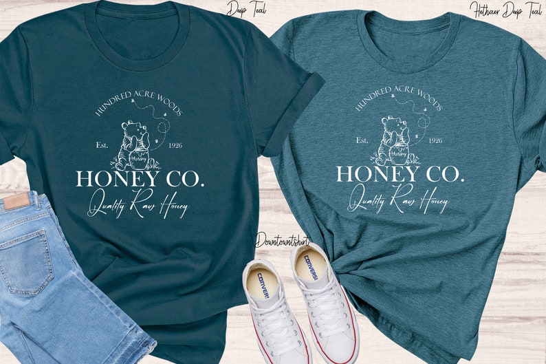 May include: Two teal blue t-shirts with a white graphic design of a bear holding a jar of honey. The graphic includes the text "Hundred Acre Woods", "Est. 1926", "Honey Co.", and "Quality Raw Honey".