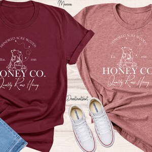May include: Two t-shirts featuring a white graphic design of a bear holding a jar of honey. The text on the shirts reads "Hundred Acre Woods" and "Honey Co. Quality Raw Honey".