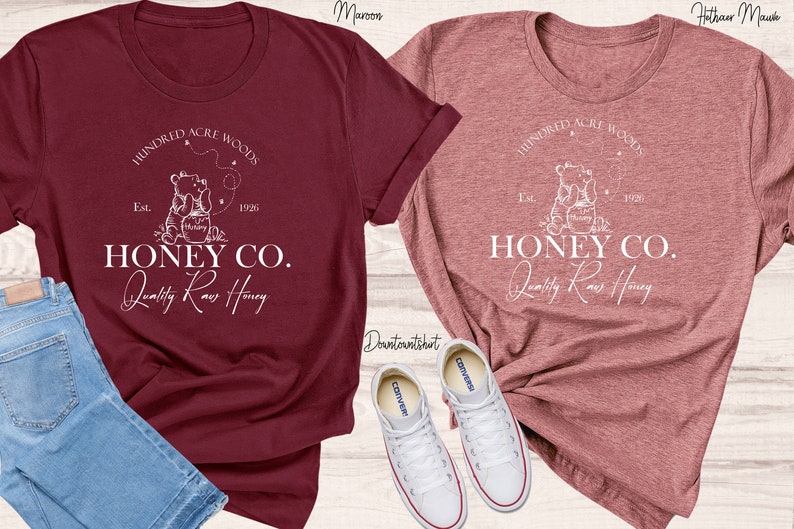May include: Two t-shirts featuring a white graphic design of a bear holding a jar of honey. The text on the shirts reads "Hundred Acre Woods" and "Honey Co. Quality Raw Honey".