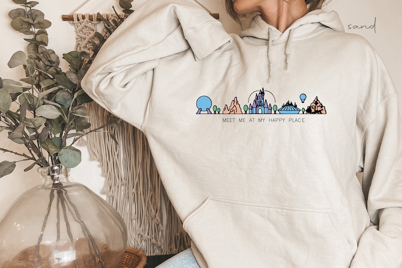 Op de afbeelding: Een lichtgrijze hoodie met een afbeelding van een gestileerde stadsgezicht en de tekst "Meet me at my happy place".