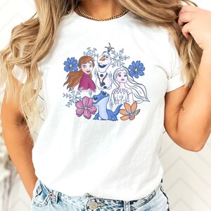 May include: White t-shirt with a colorful graphic print of characters from the movie Frozen. The characters include Elsa, Anna, Olaf, and a blue lizard. The graphic is surrounded by snowflakes and flowers.