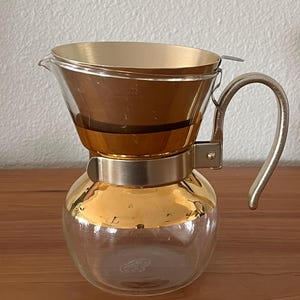 May include: A clear glass coffee maker with a gold-colored metal filter and handle. The coffee maker has a round base and a conical top, with a gold-colored metal band around the middle. The handle is curved and made of the same gold-colored metal.