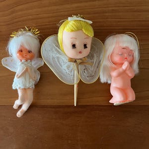 May include: Three vintage angel figurines. One angel is dressed in a white dress with gold wings and a halo. Another angel has a yellow head with a white halo and a white lace collar. The third angel is nude with white hair and praying hands.