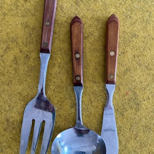 May include: A stainless steel fork, ladle, and spreader knife set. Each utensil has a wooden handle with a brass rivet. The handles are a dark brown color. The utensils are arranged on a yellow surface.