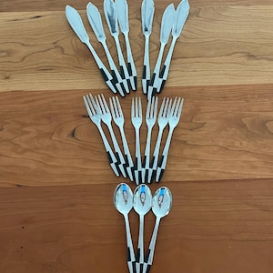 May include: A collection of stainless steel silverware, including butter knives, forks, and spoons. The handles feature a black and white checkered design. The silverware is arranged on a wooden surface.