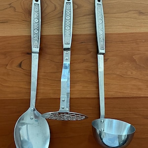 May include: Three stainless steel kitchen utensils: a serving spoon, a potato masher, and a ladle. Each utensil has a decorative floral pattern on the handle. The spoon and ladle have rounded bowls, while the masher has a flat, perforated head.